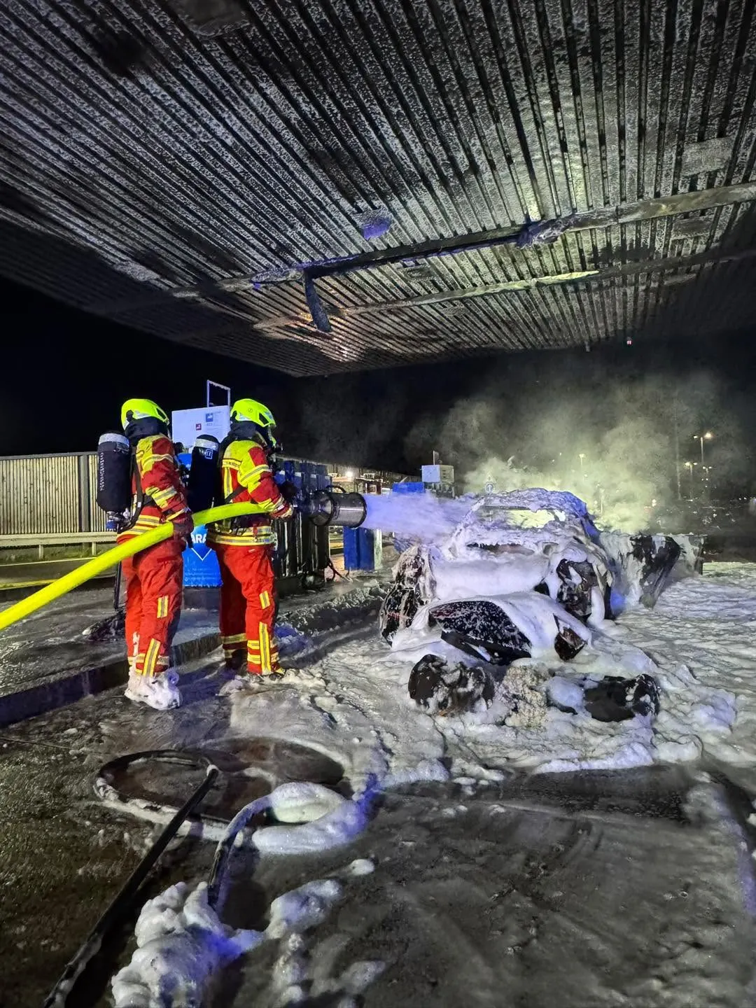 Zwei Feuerwehrleute in Schutzkleidung löschen ein stark rauchendes, brennendes Fahrzeug an einer Tankstelle mit Schaum.