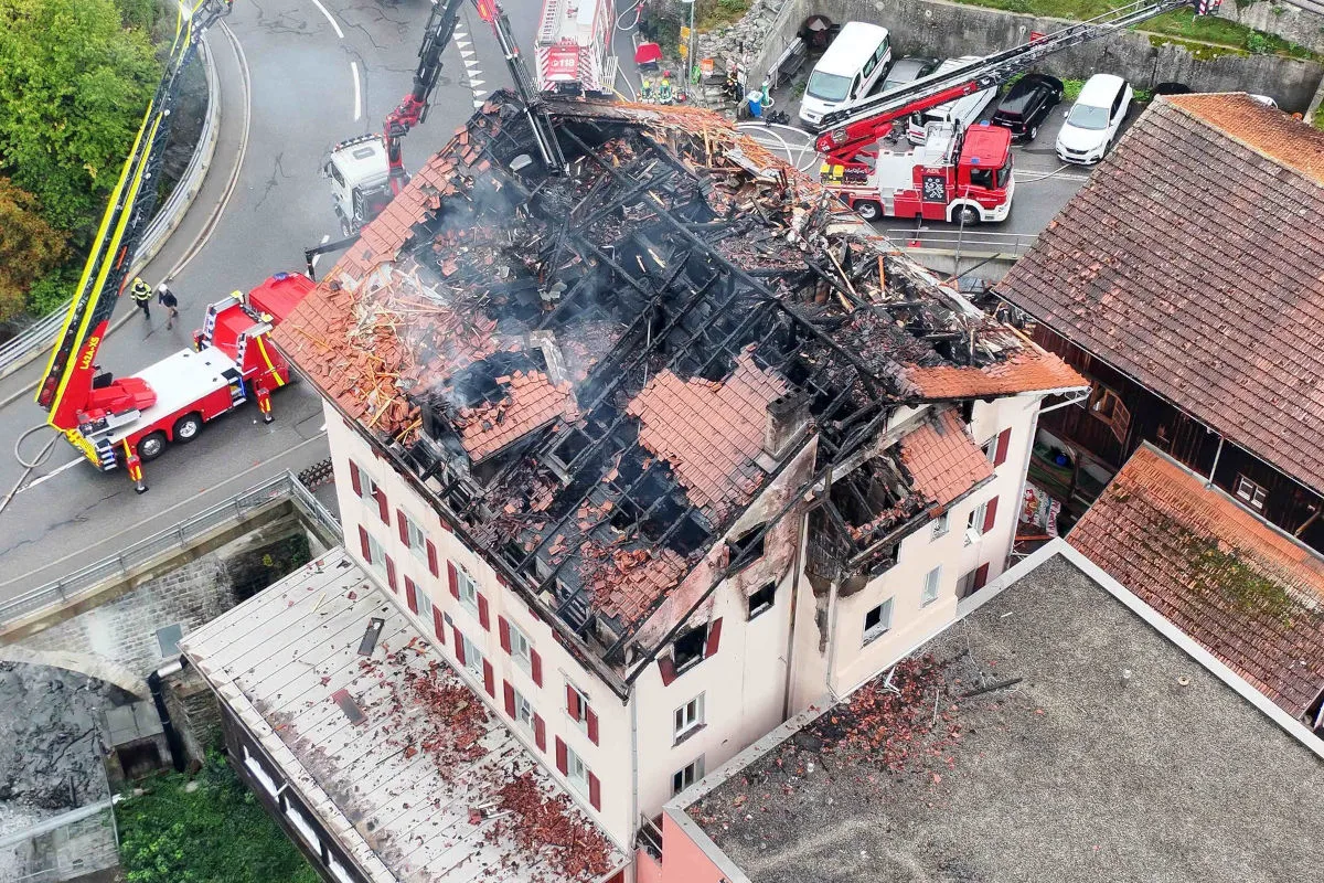 Das Bild zeigt ein schwer beschädigtes Dach eines Hauses, das nach einem Brand verkohlt ist. Feuerwehrfahrzeuge und Einsatzkräfte sind vor Ort sichtbar, um den Brand zu bekämpfen.