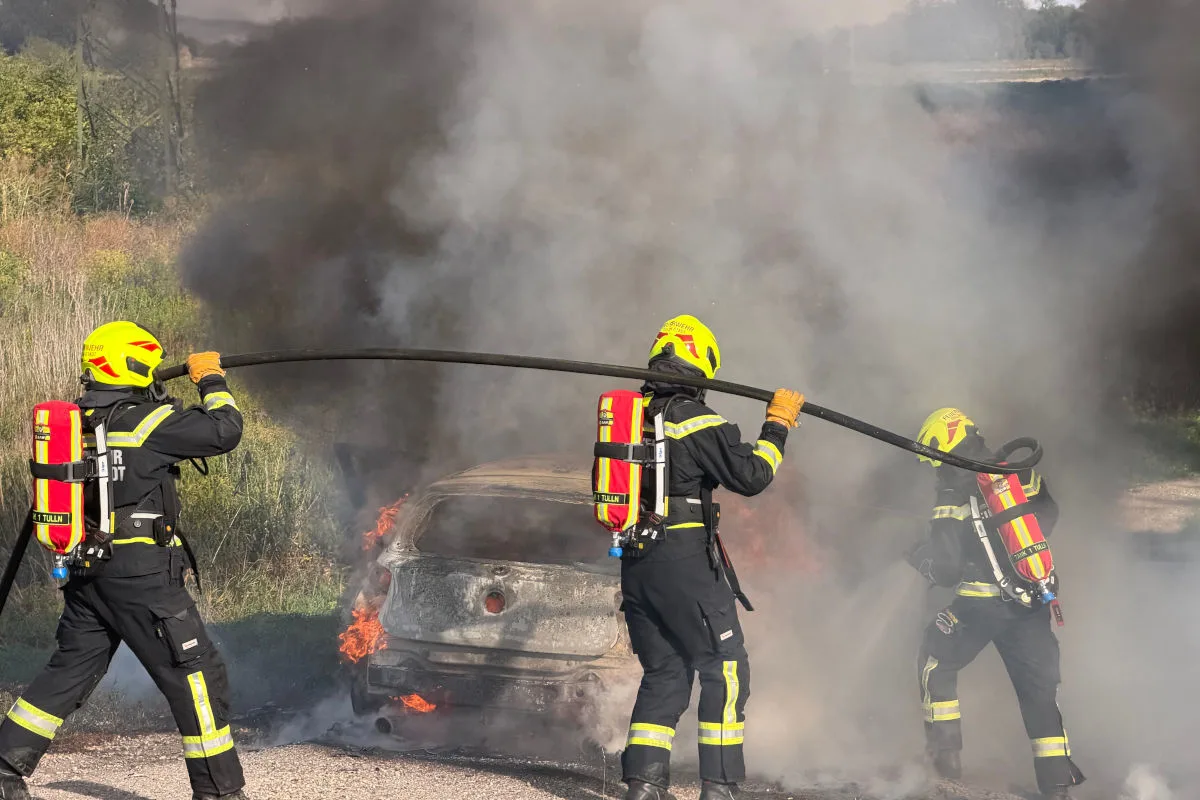 Drei Feuerwehrleute in voller Schutzausrüstung verwenden einen Schlauch, um ein Auto zu löschen, das in Flammen steht und von Rauch umgeben ist.