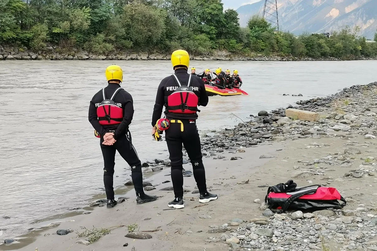 Zwei Feuerwehrleute in Schutzkleidung stehen am Flussufer und beobachten ein mit mehreren Personen besetztes Schlauchboot im Wasser. Der Bereich ist von Bäumen umgeben, und im Hintergrund sind Berge zu sehen.