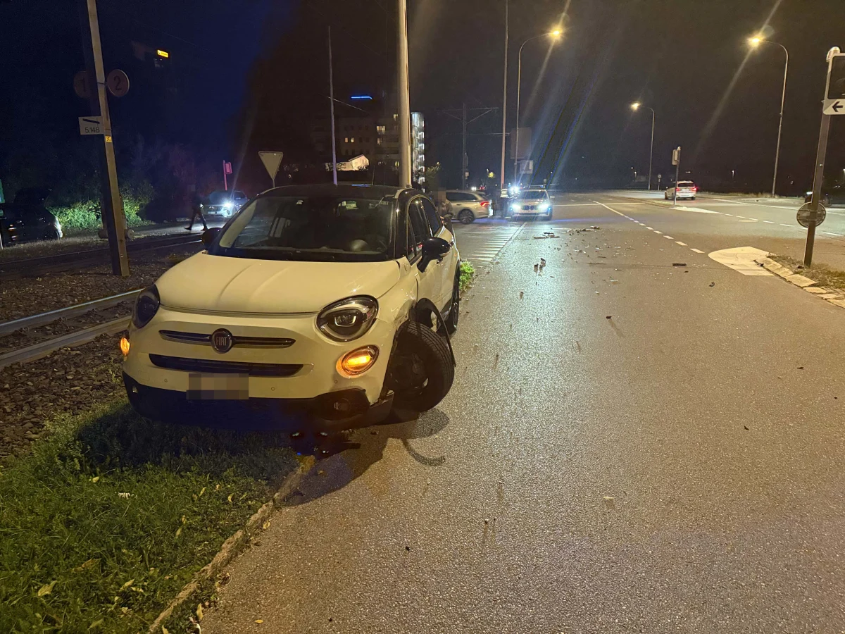 Ein weißer Fiat steht mit einem abgerissenen Rad auf dem Bordstein neben einer Straße bei Nacht. Im Hintergrund sind weitere Fahrzeuge sichtbar.