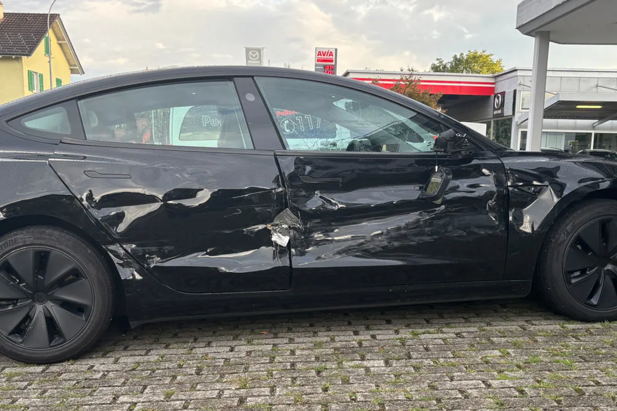 Ein schwarzer Tesla mit stark deformierter Seite steht auf einem Parkplatz. Im Hintergrund ist eine Tankstelle zu sehen.
