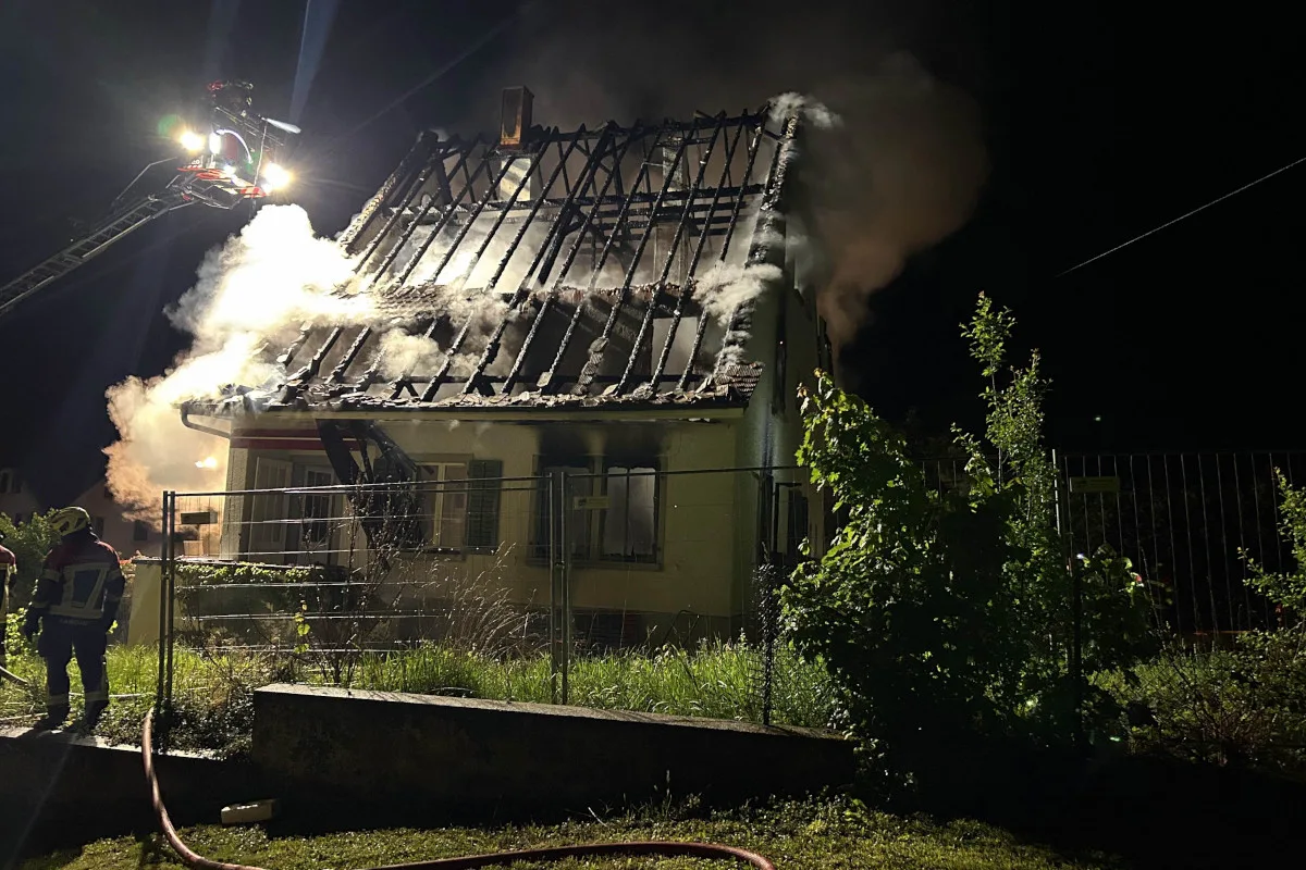Bei Nacht löscht die Feuerwehr ein brennendes Haus. Rauch steigt vom Dach auf, während Feuerwehrleute arbeiten.