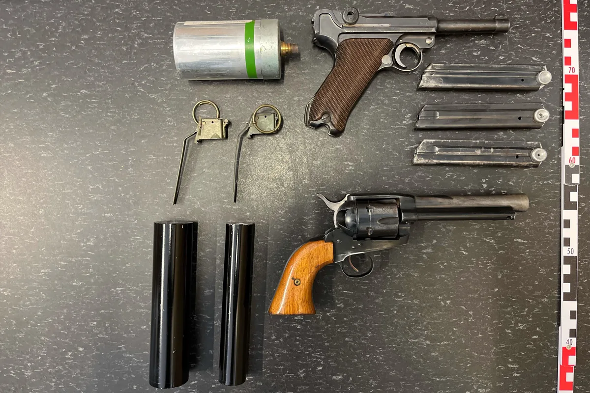 Zwei Pistolen, mehrere Magazine, eine Metalldose und zwei Metallringe auf einer schwarzen Oberfläche fotografiert. Rechts ist ein Maßstab sichtbar.