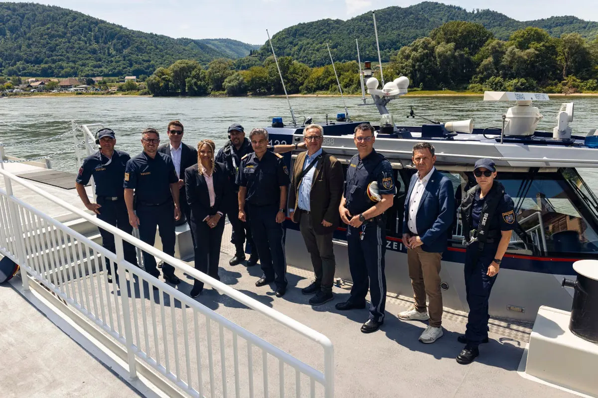 Mehrere Personen, darunter Polizeiangehörige und Zivilisten, posieren vor einem Polizeiboot an einem Fluss, umrahmt von bewaldeten Hügeln im Hintergrund.