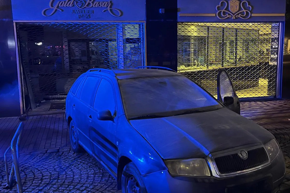 Ein blauer Pkw steht nachts vor einem Juweliergeschäft. Das metallene Sicherheitsgitter der Eingangstür ist geöffnet.