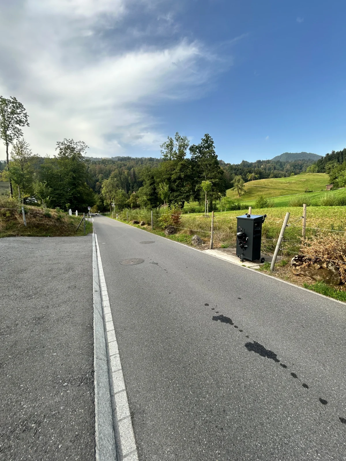 Eine Landstraße führt durch eine grüne, ländliche Gegend. Auf der Straße ist eine dunkle Spur sichtbar, die möglicherweise Öl ist. Der Himmel ist teilweise bewölkt und die Umgebung ist ruhig und ländlich.