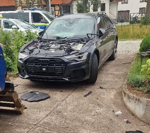 Ein schwarzer Audi mit beschädigter Front und geöffneter Motorhaube steht auf einer Auffahrt; im Hintergrund ist ein Polizeiwagen zu sehen.