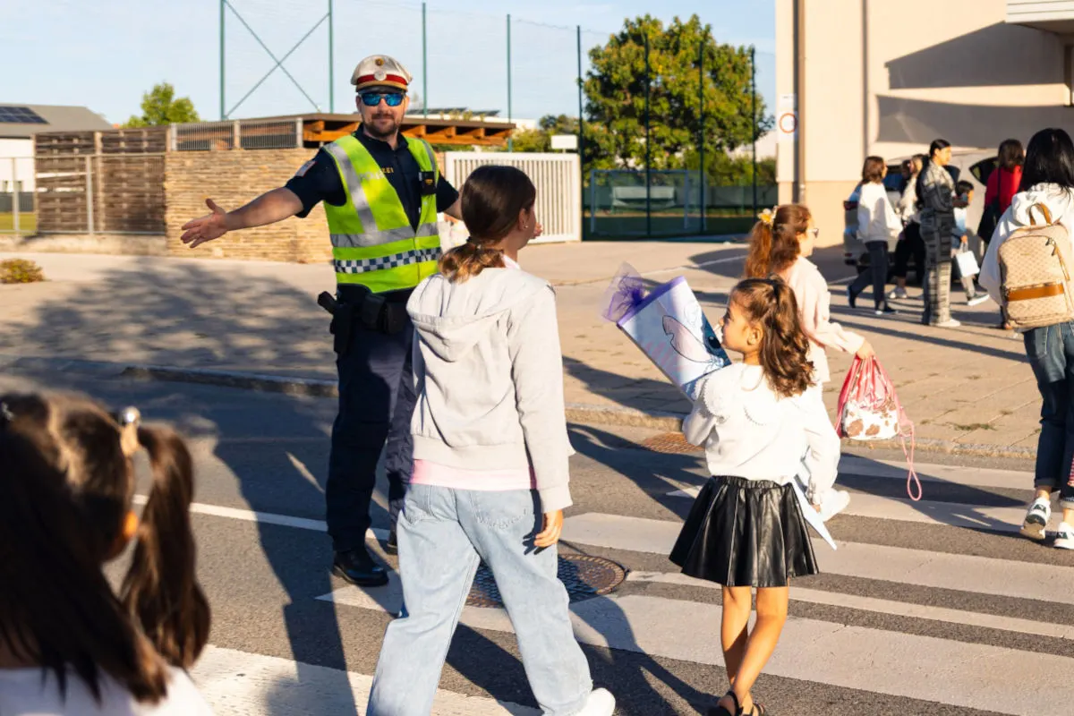Ein Polizist in Warnweste assistiert einer Gruppe von Kindern auf einem Zebrastreifen. Eines der Kinder hält eine Schultüte.