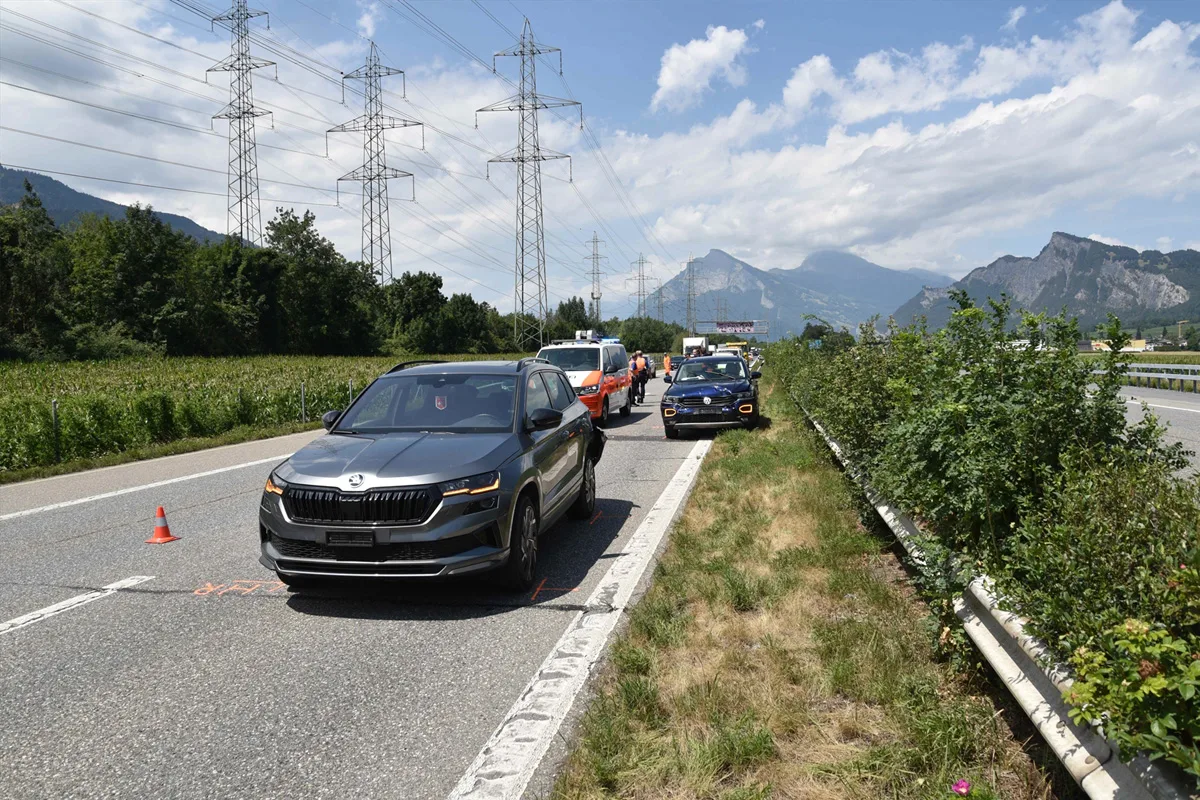 Mehrere Autos und ein Van stehen auf einer zweispurigen Landstraße in bergiger Umgebung. Stromleitungen verlaufen parallel zur Straße.