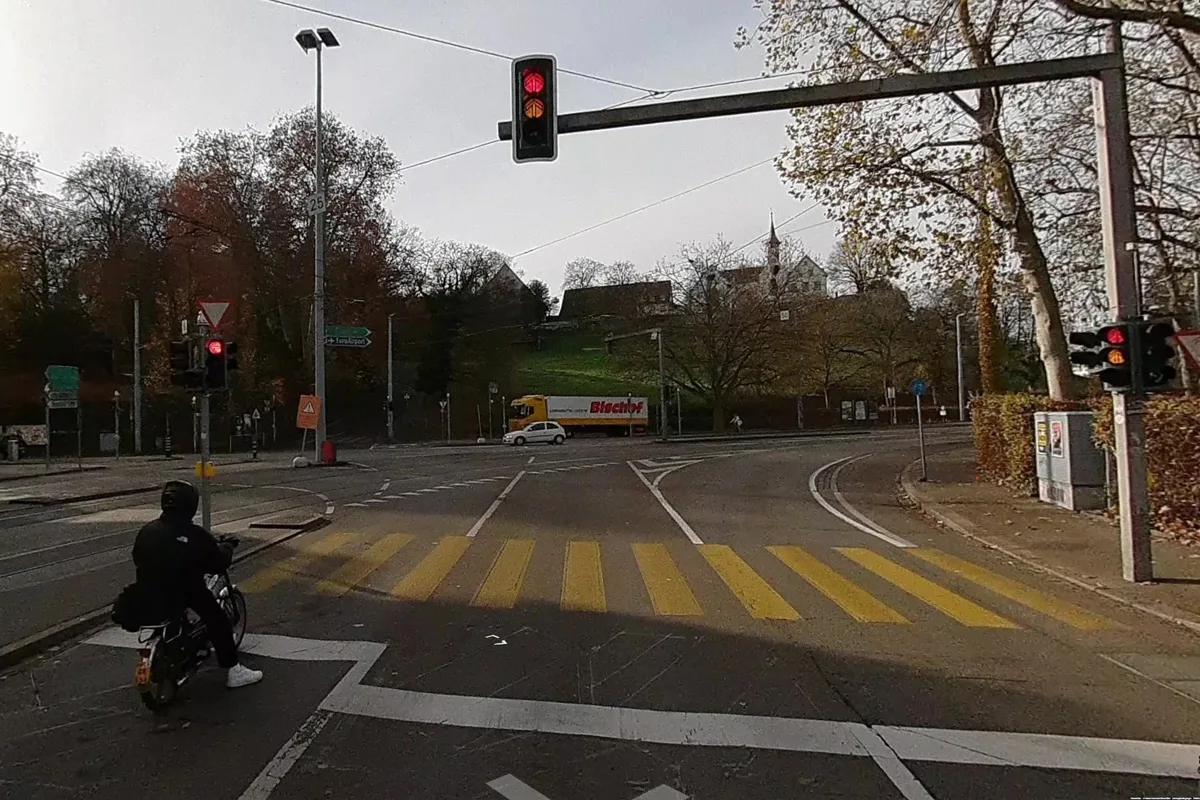 Ein Motorradfahrer wartet an einer roten Ampel vor einer Kreuzung mit einem Lkw im Hintergrund. Die Szene zeigt eine herbstliche Umgebung.