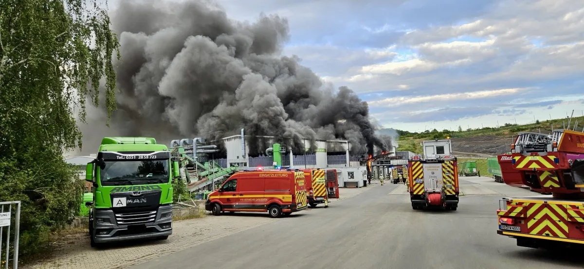 Dichte schwarze Rauchwolken steigen von einer Industrieanlage auf, umgeben von mehreren Feuerwehrfahrzeugen. Ein grüner Lastwagen steht am Rand.