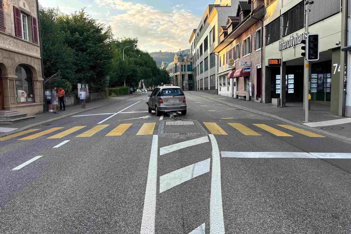 Ein graues Auto steht auf einem Zebrastreifen in einer städtischen Straße mit umliegenden Gebäuden und Bäumen.