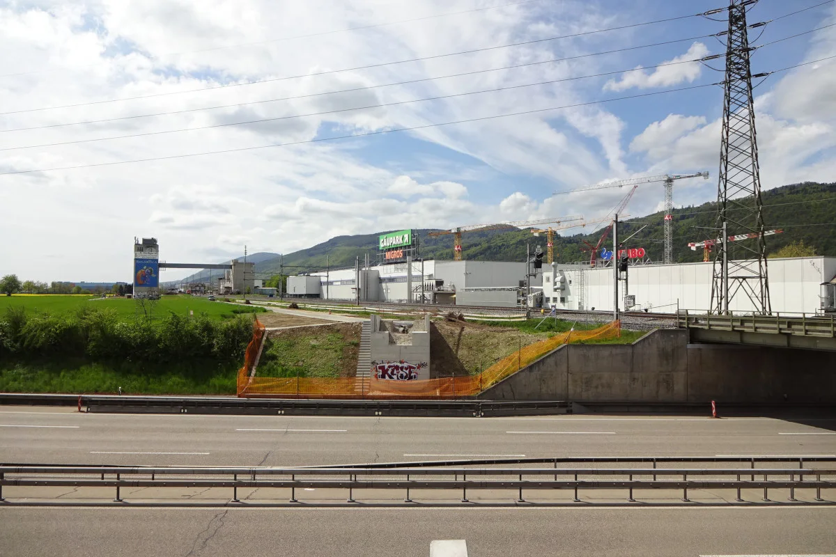 Das Bild zeigt eine leere Autobahn im Vordergrund und ein Industriegebiet im Hintergrund mit mehreren Kränen. Ein grüner Hügel ragt im Hintergrund empor, während der Himmel teils bewölkt ist.