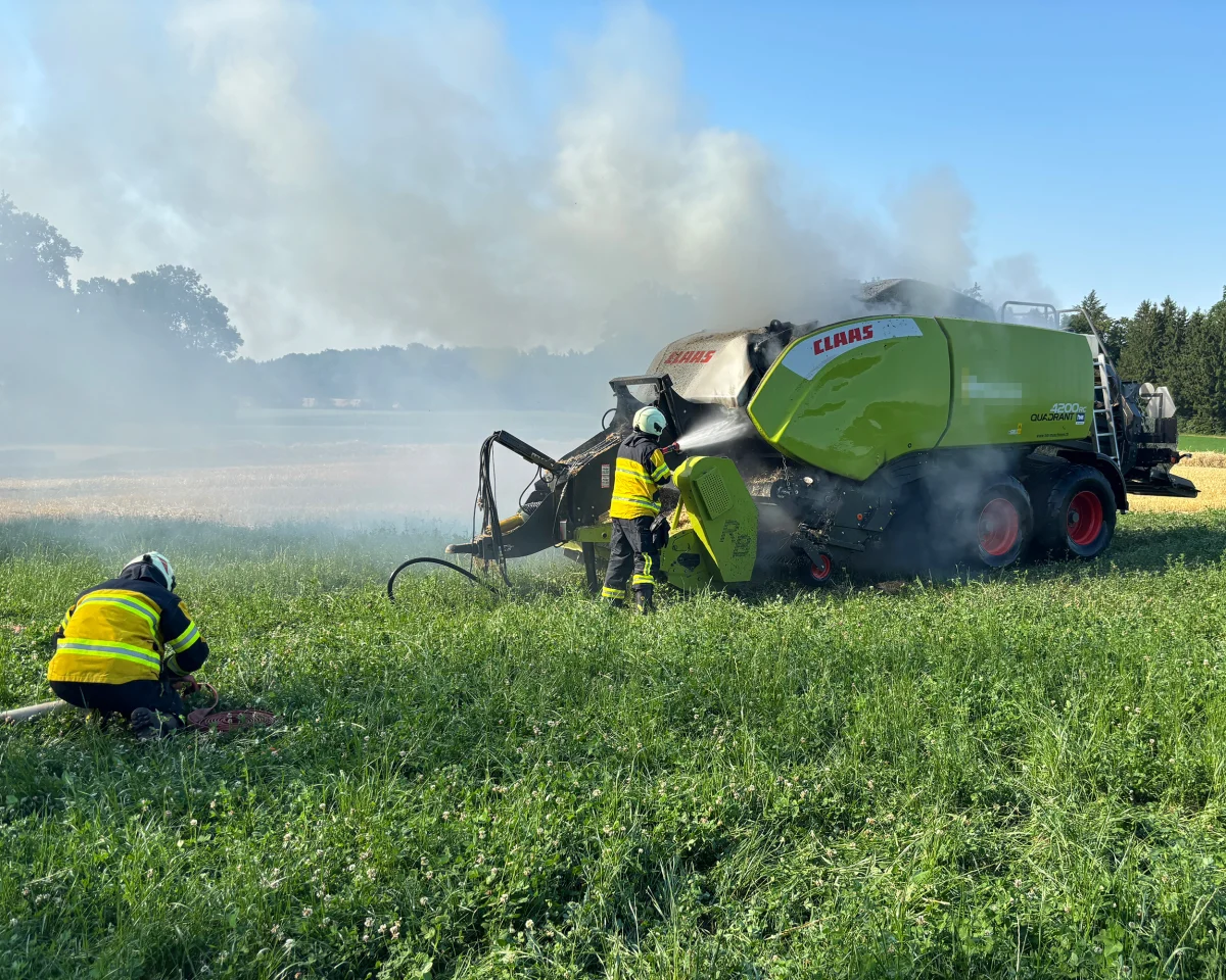Eine Ballenpresse, die auf einem Feld raucht, wird von Feuerwehrleuten gelöscht. Dichte Rauchwolken steigen auf.