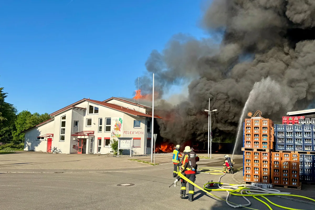 Mehrere Feuerwehrleute bekämpfen einen massiven Brand in einem großen Gebäude, dichter schwarzer Rauch steigt auf.
