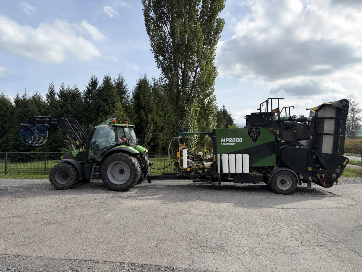 Ein grüner Traktor ist mit einem MP2000-Kompaktierer verbunden und steht auf einer asphaltierten Straße. Im Hintergrund sind Bäume und ein bewölkter Himmel zu sehen.