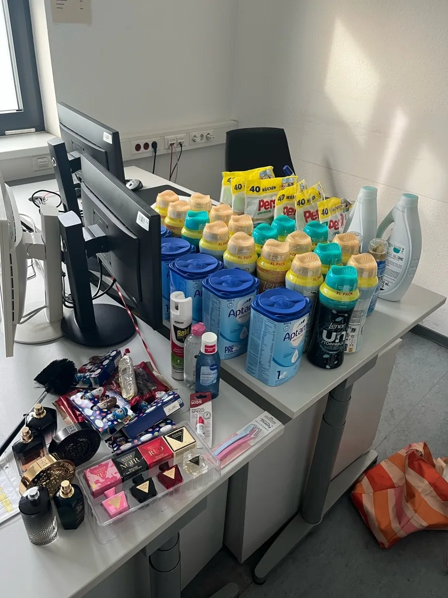 Auf einem Tisch stehen gestohlene Waren, darunter Verpackungen von Babynahrung, Waschmittel und Kosmetika. Im Hintergrund sind Computerbildschirme zu sehen.