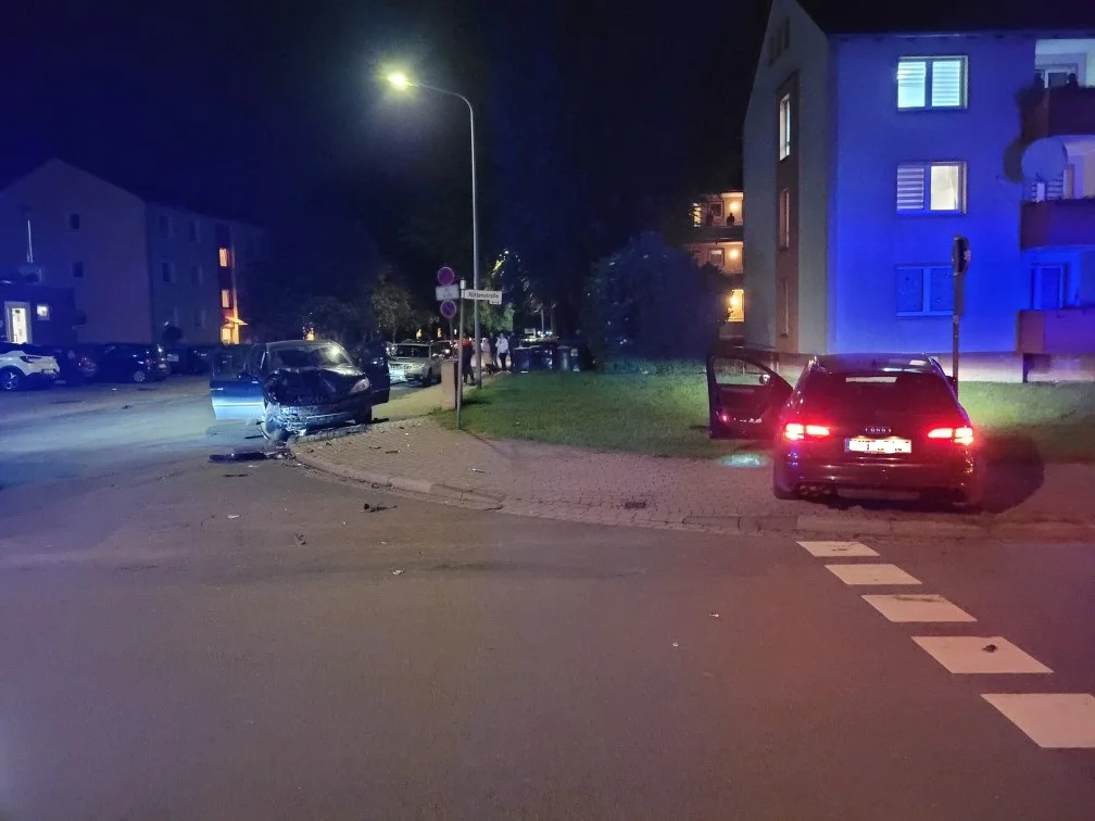 Bei Nacht sind zwei stark beschädigte Autos an einer Kreuzung zu sehen. Eins der Fahrzeuge steht auf dem Gehweg, während das andere im Straßenbereich steht.