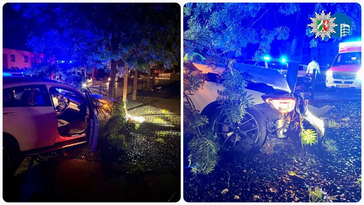 Ein weißes Auto ist frontal gegen einen Baum geprallt. Im Hintergrund sind Rettungsfahrzeuge mit Blaulicht zu sehen.
