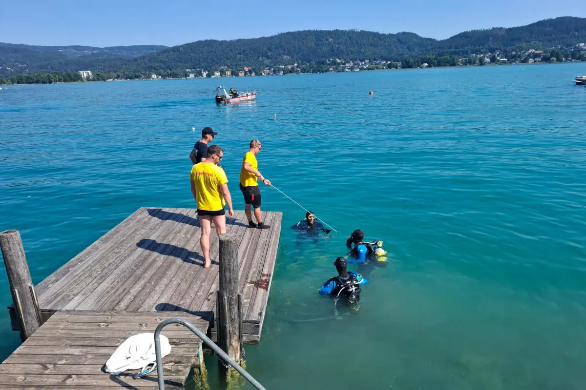 Mehrere Einsatzkräfte stehen auf einem Steg am See, während Taucher im Wasser aktiv sind und ein Boot im Hintergrund sichtbar ist.