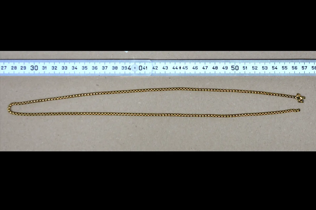 Eine goldene Kette ist auf einem braunen Hintergrund ausgebreitet. Direkt darüber liegt ein Lineal, das Größe und Länge der Kette verdeutlicht.