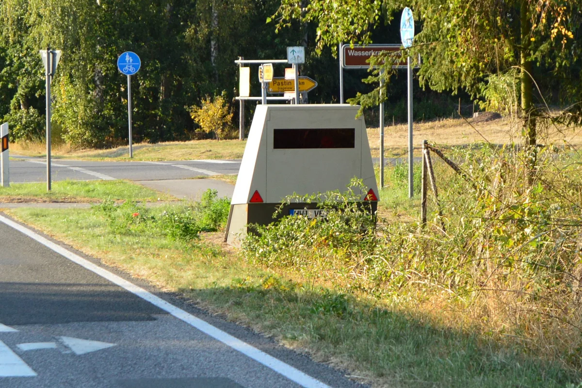Ein mobiler Blitzer steht gut getarnt am Straßenrand einer Landstraße. Verkehrszeichen und Vegetation sind im Hintergrund sichtbar.