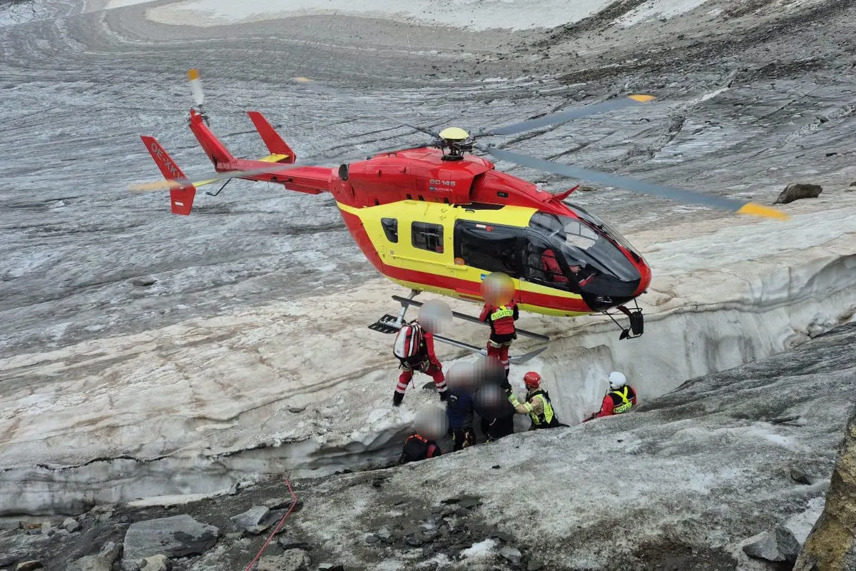 Ein rot-gelber Rettungshubschrauber schwebt über einem Gletscher, während Einsatzkräfte Personen aus einer Spalte bergen.