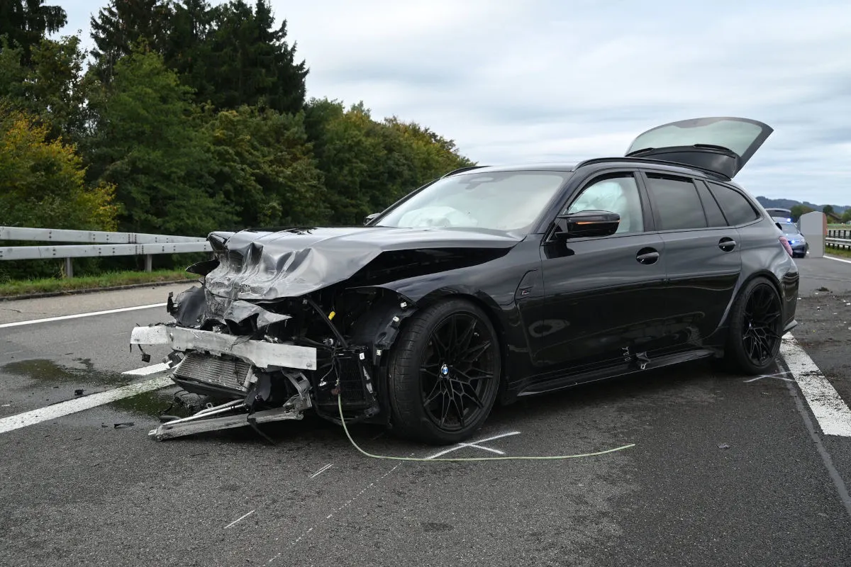 Ein stark beschädigter schwarzer BMW mit offenem Kofferraum steht auf einer Landstraße mit Schutzplanken im Hintergrund.