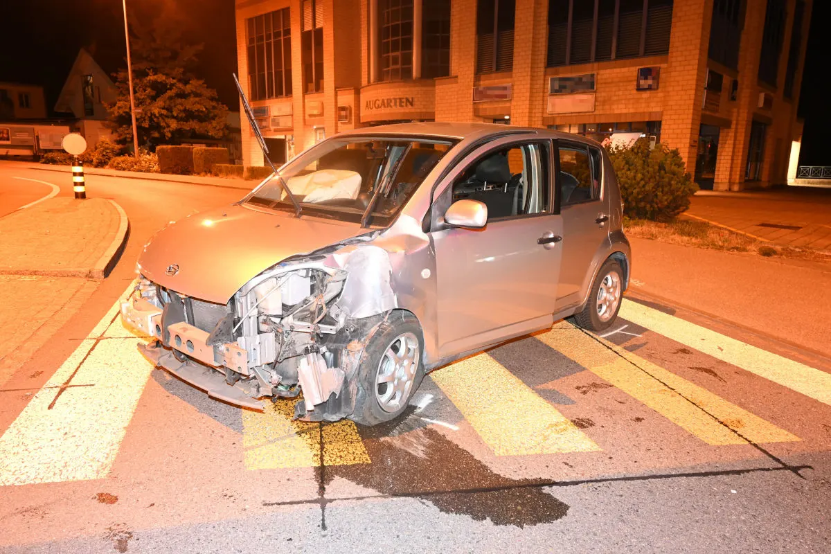 Ein Auto mit einem erheblichen Frontschaden steht auf einem beleuchteten Zebrastreifen vor einem Gebäude. Der Schaden deutet auf eine Kollision hin.