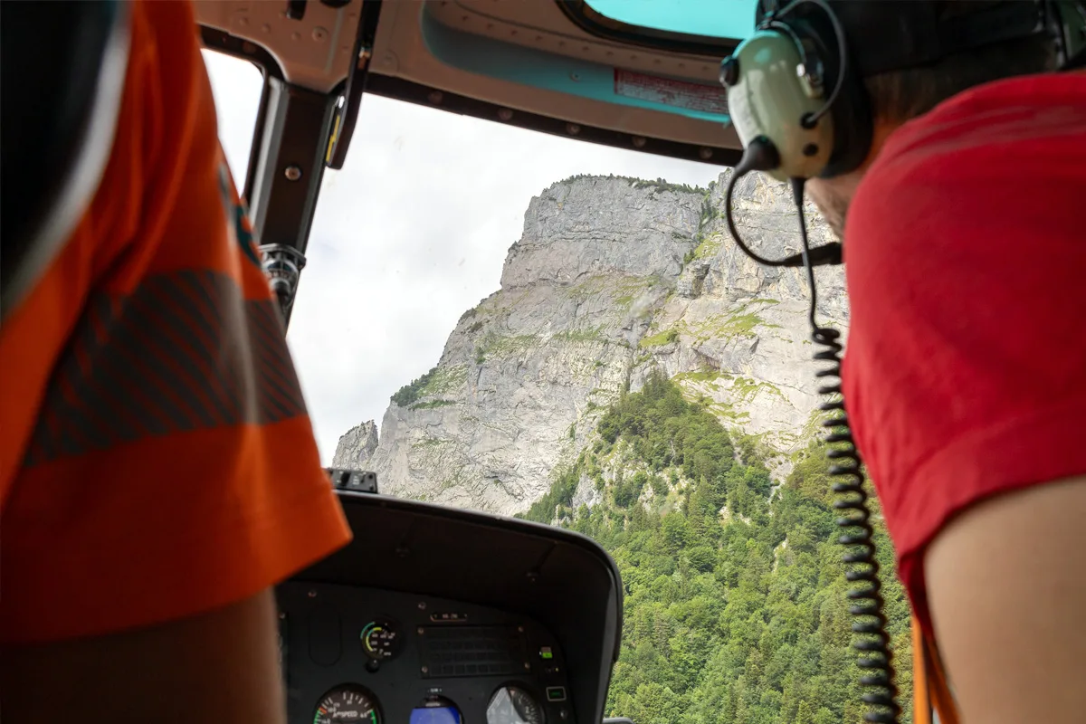 Der Blick aus einem Hubschrauber führt über ein Armaturenbrett hin zu einer felsigen Berglandschaft. Links im Bild ist eine Person in orangener Kleidung zu sehen.