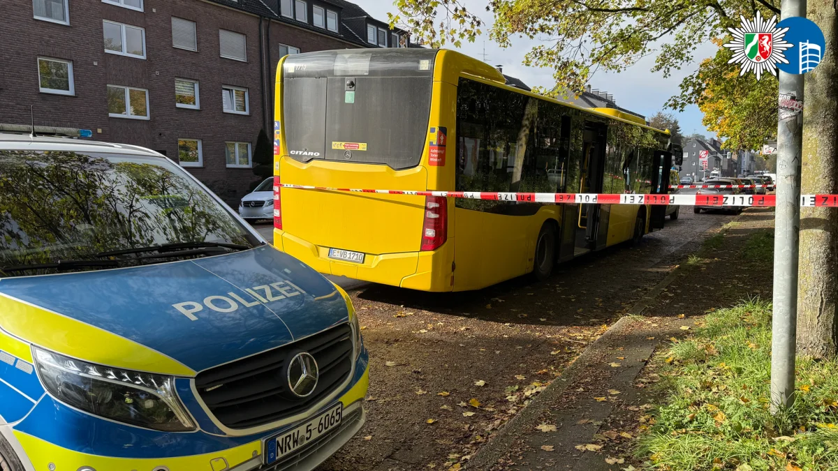 Ein Polizeiwagen und ein gelber Linienbus stehen hinter einem Absperrband an einer Straßenkreuzung in einem Wohngebiet. Herbstliches Laub bedeckt den Boden.