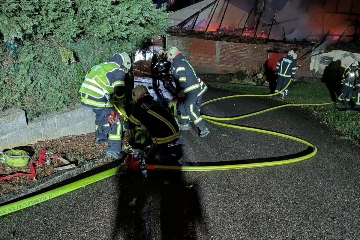 Mehrere Feuerwehrleute in Schutzkleidung sind vor einem brennenden Gebäude im Einsatz. Schläuche liegen auf dem Boden, während das Feuer im Hintergrund sichtbar ist.