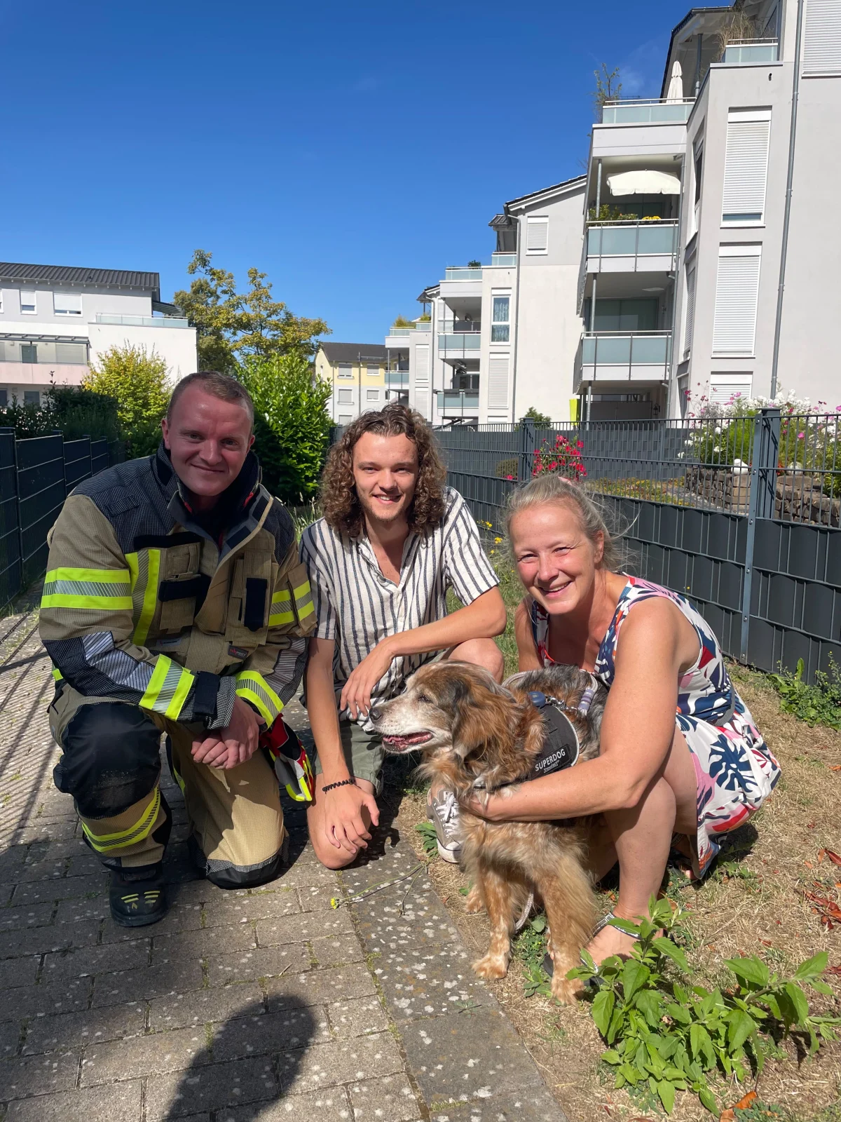 Ein Feuerwehrmann in Uniform kniet draußen neben zwei Personen und einem Hund. Im Hintergrund sind Wohngebäude zu sehen.