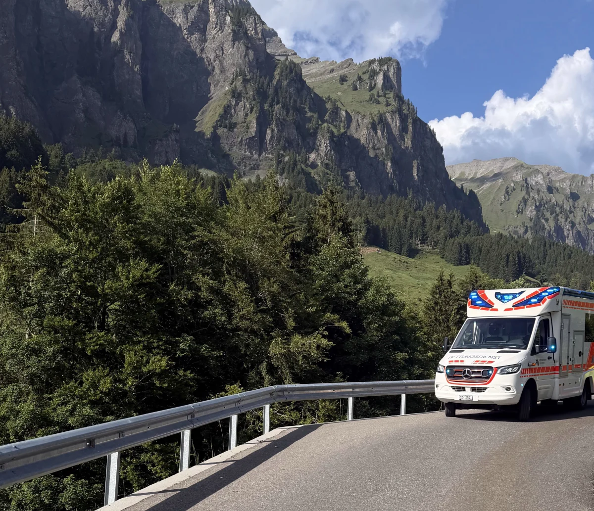 Ein Rettungswagen der Marke Mercedes fährt auf einer kurvigen Bergstraße. Im Hintergrund sind hohe Felsformationen und bewaldete Hänge zu sehen.