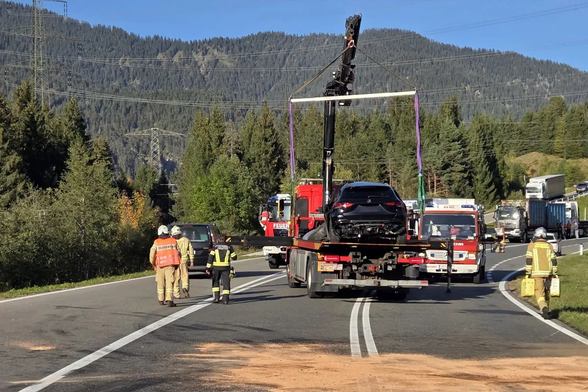 Ein schwarzes Auto wird auf einer Bergstraße mithilfe eines Krans auf einen Transporter gehoben. Feuerwehrleute sichern die Unfallstelle ab.