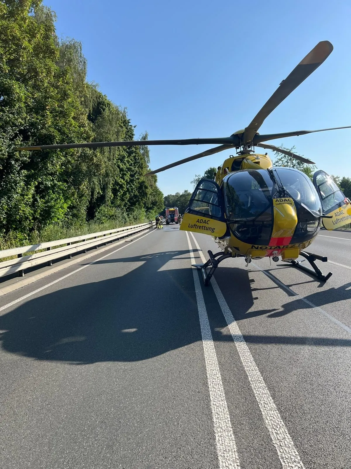 Ein ADAC Rettungshubschrauber steht auf einer zweispurigen Straße, während im Hintergrund Bäume zu sehen sind. Es ist ein klarer Tag.