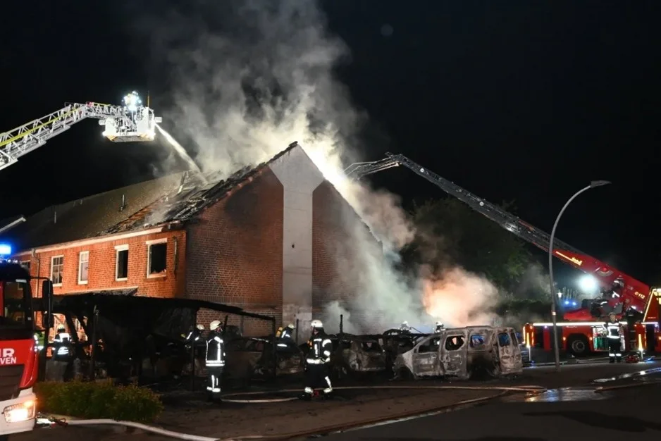 Feuerwehrkräfte bekämpfen bei Nacht einen Großbrand in einem einstöckigen Gebäude, Rauch steigt aus dem Dach.