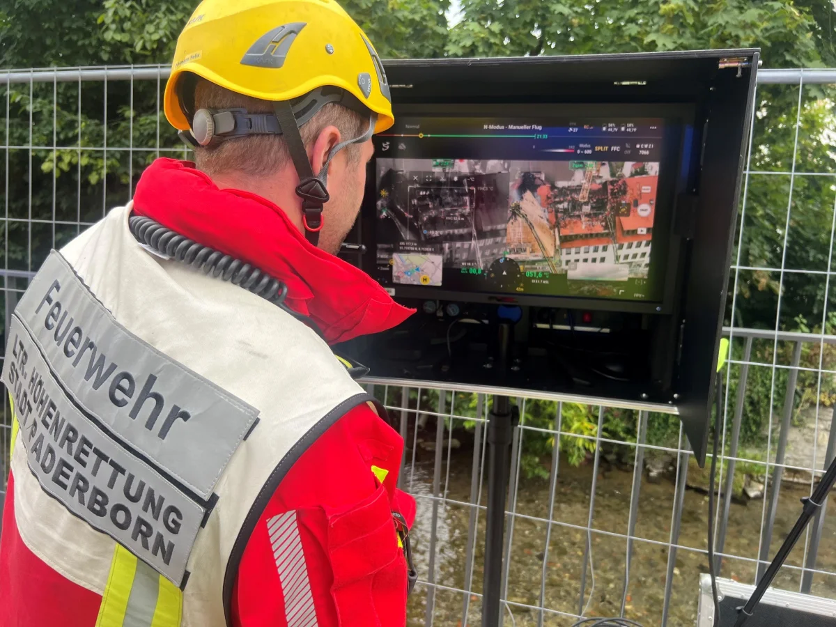 Ein Feuerwehrmann der Höhenrettung Paderborn schaut auf einen Monitor, der Luftbilder zeigt. Eine Absperrung und Bäume sind im Hintergrund sichtbar.