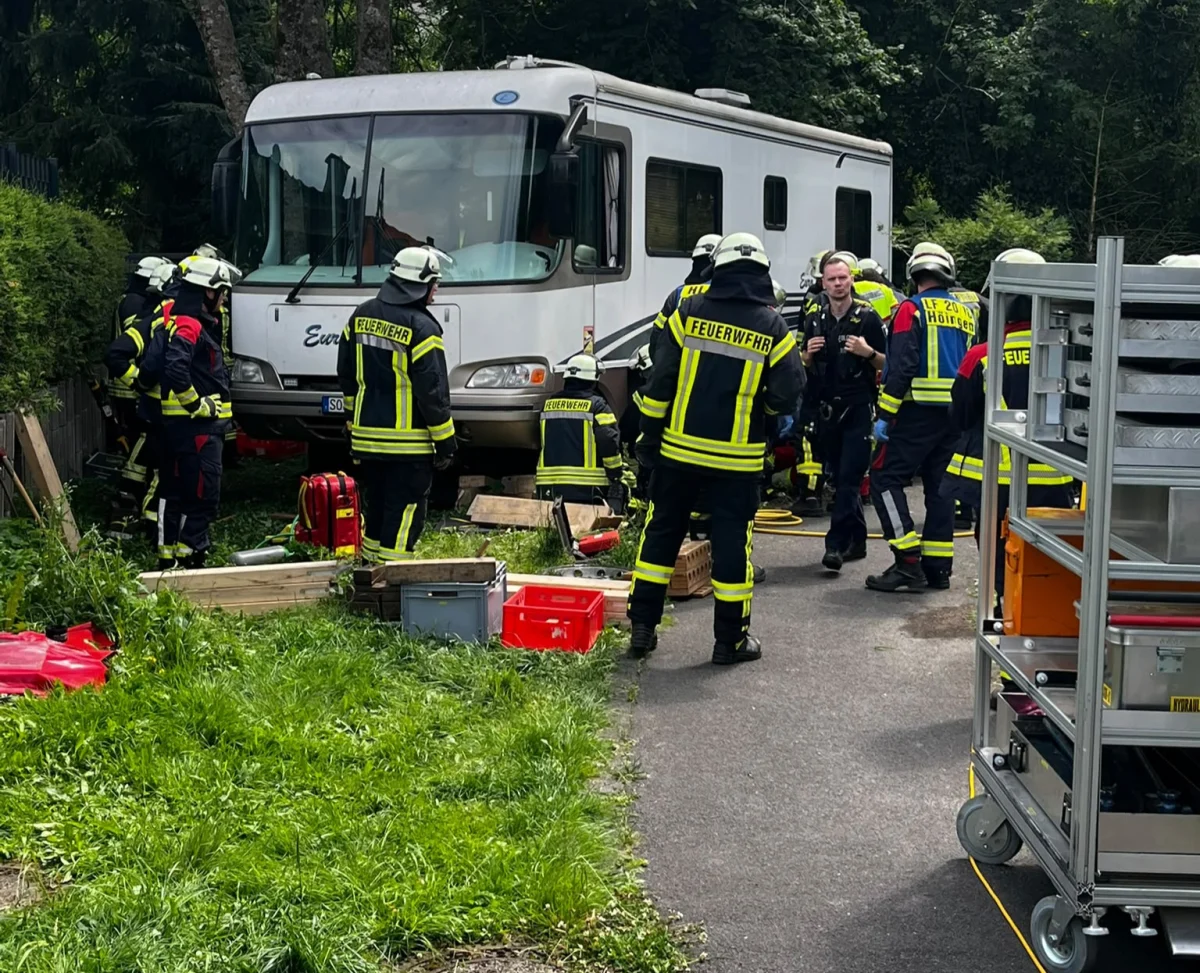 Mehrere Feuerwehrleute arbeiten an einem Wohnmobil, das im Grünen geparkt ist. Es sind verschiedene Ausrüstungsgegenstände um das Fahrzeug verteilt.