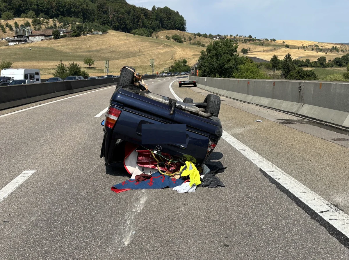 Ein Auto liegt umgekippt auf dem Dach in der Mitte einer Autobahn. Die Umgebung wirkt ländlich mit Feldern und Hügeln.