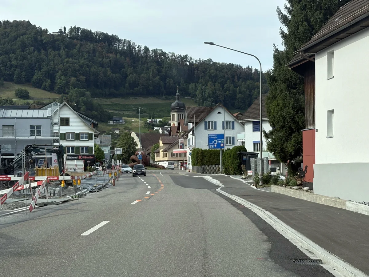 Eine Dorfstraße mit laufenden Straßenarbeiten, umgeben von Wohnhäusern. Im Hintergrund ist eine Kirche zu sehen.