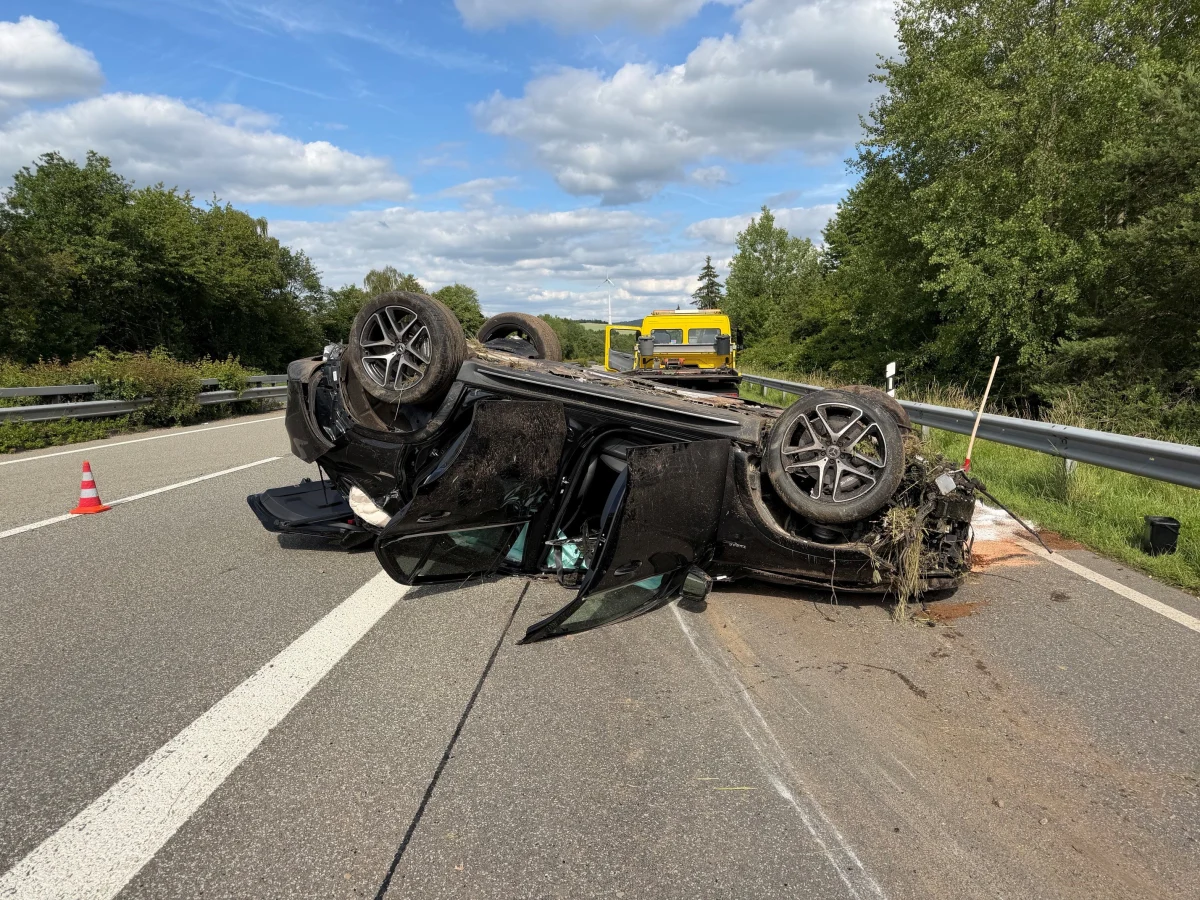 Das Bild zeigt ein Auto, das auf einer Autobahn auf dem Dach liegt. Ein Verkehrskegel steht daneben und ein Abschleppwagen ist im Hintergrund sichtbar.