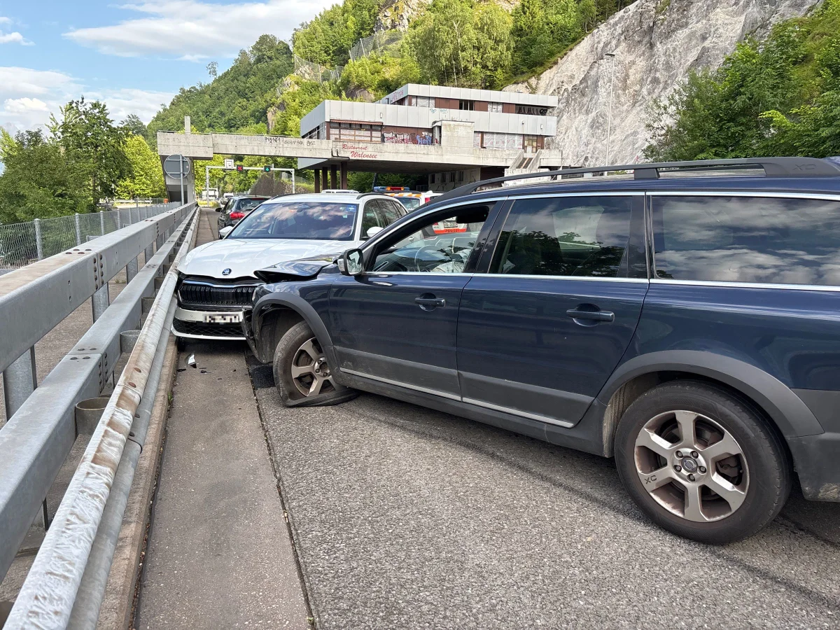 Zwei Fahrzeuge sind sichtbar, die nach einem Zusammenstoß an einer Leitplanke stehen. Im Hintergrund befindet sich ein Gebäude und bewaldetes Gelände.