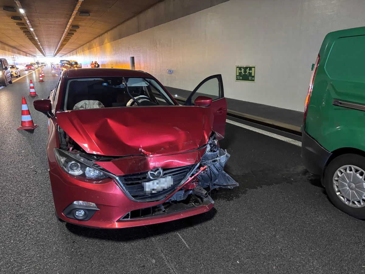 Ein rotes Auto mit erheblichen Frontschäden steht in einem Tunnel. Im Hintergrund sind weitere Fahrzeuge und Verkehrskegel sichtbar.
