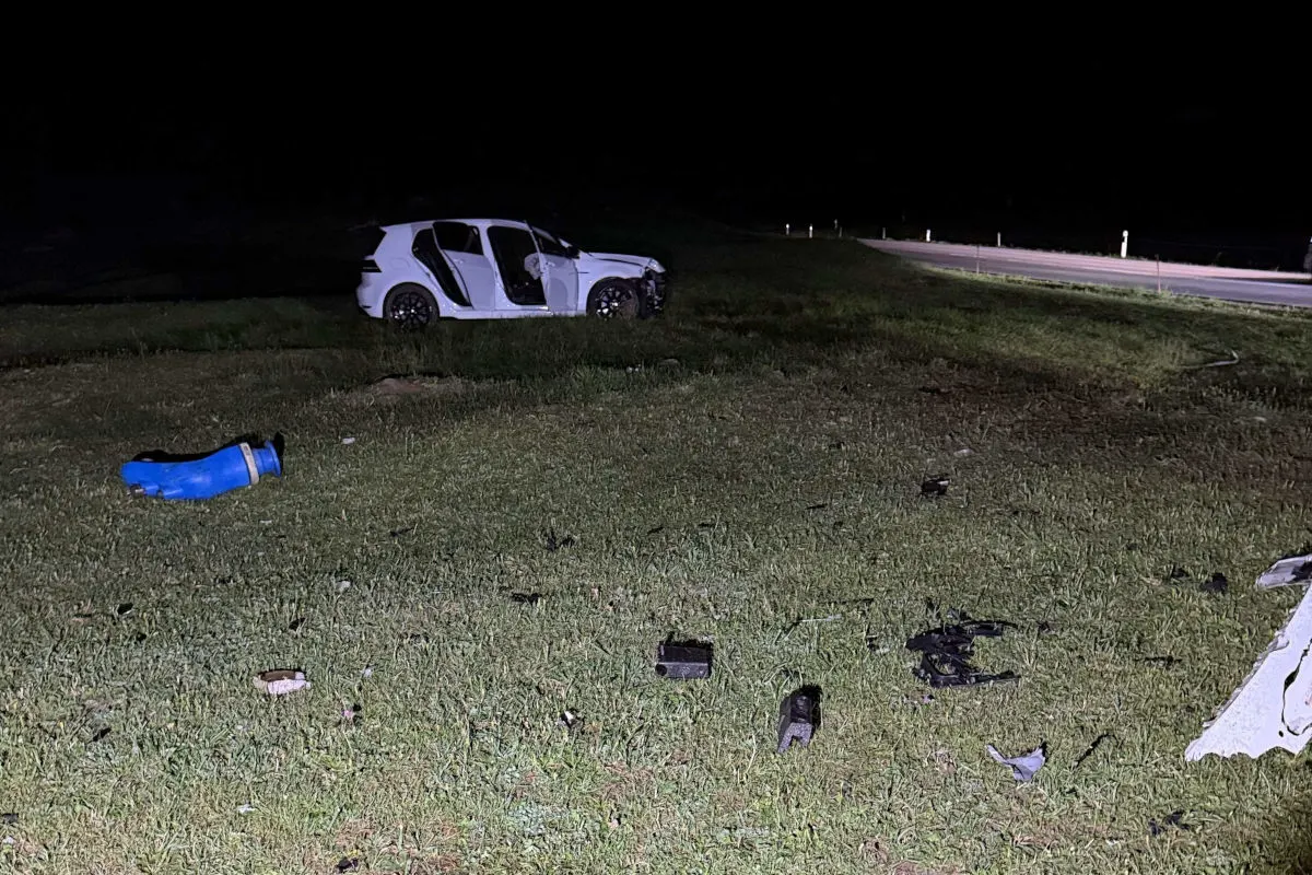 Ein weißes Auto mit offen stehender Tür steht beschädigt auf einer Wiese bei Nacht. Um das Fahrzeug herum liegen Trümmerteile verstreut.