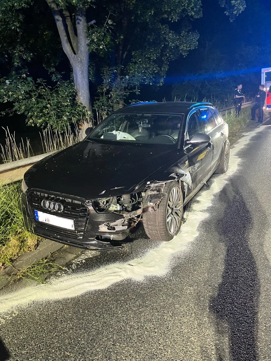Ein schwarzer Audi mit sichtbaren Schäden an der Front steht bei Nacht am Straßenrand. Der Bereich ist abgesperrt.