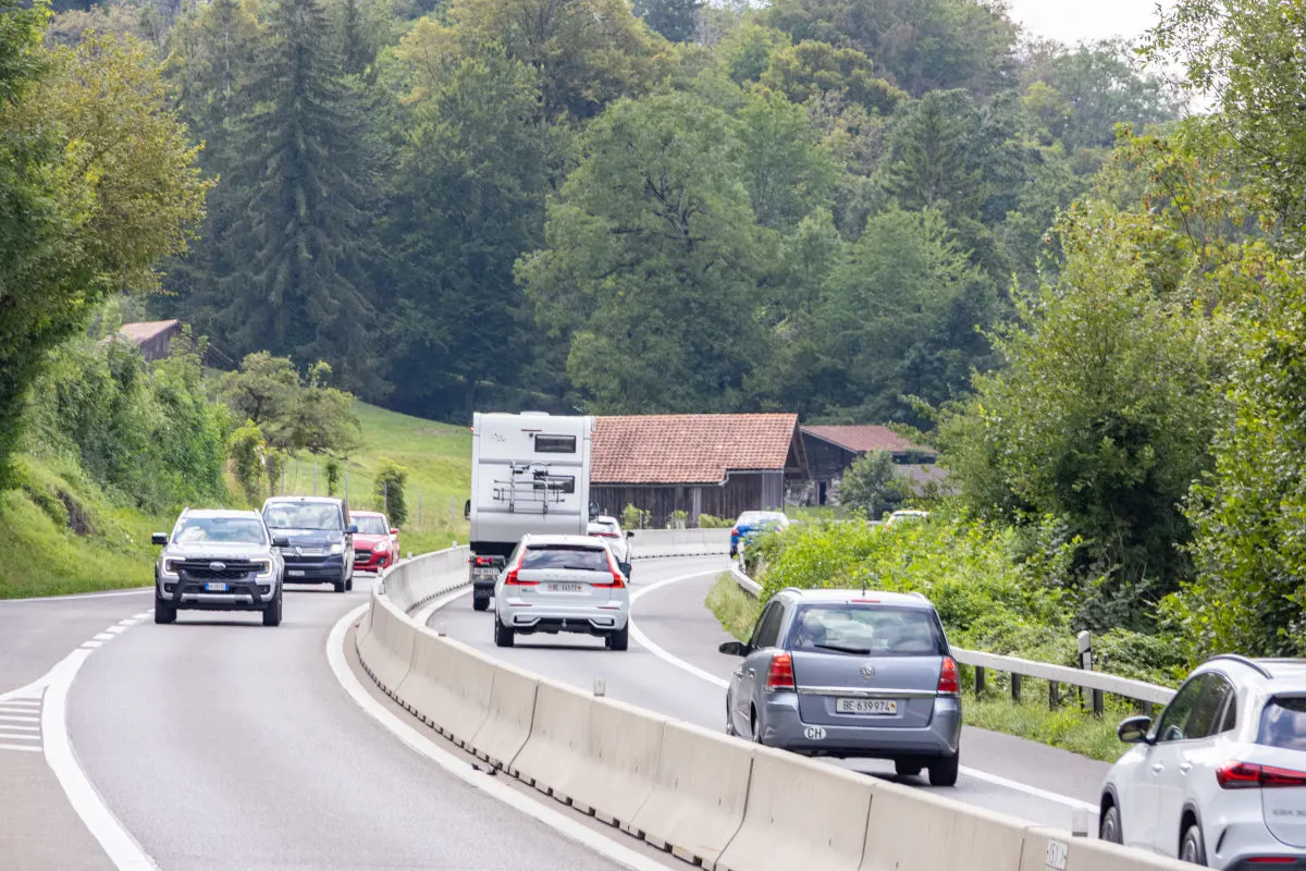Verschiedene Fahrzeuge fahren auf einer kurvigen Landstraße, umgeben von grüner Landschaft und Wäldern.