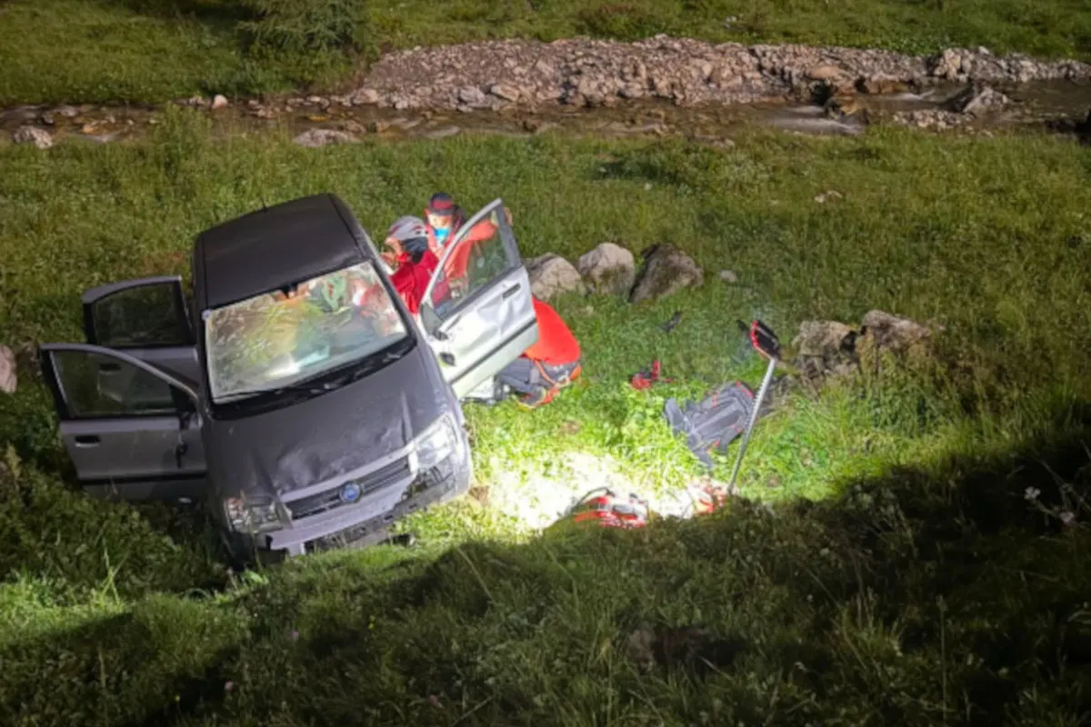 Ein graues Auto steht bei Nacht in einer Wiese nahe einem Bach, umgeben von Rettungskräften im Einsatz. Die Türen des Fahrzeugs sind geöffnet und Einsatzmaterial liegt auf dem Boden.