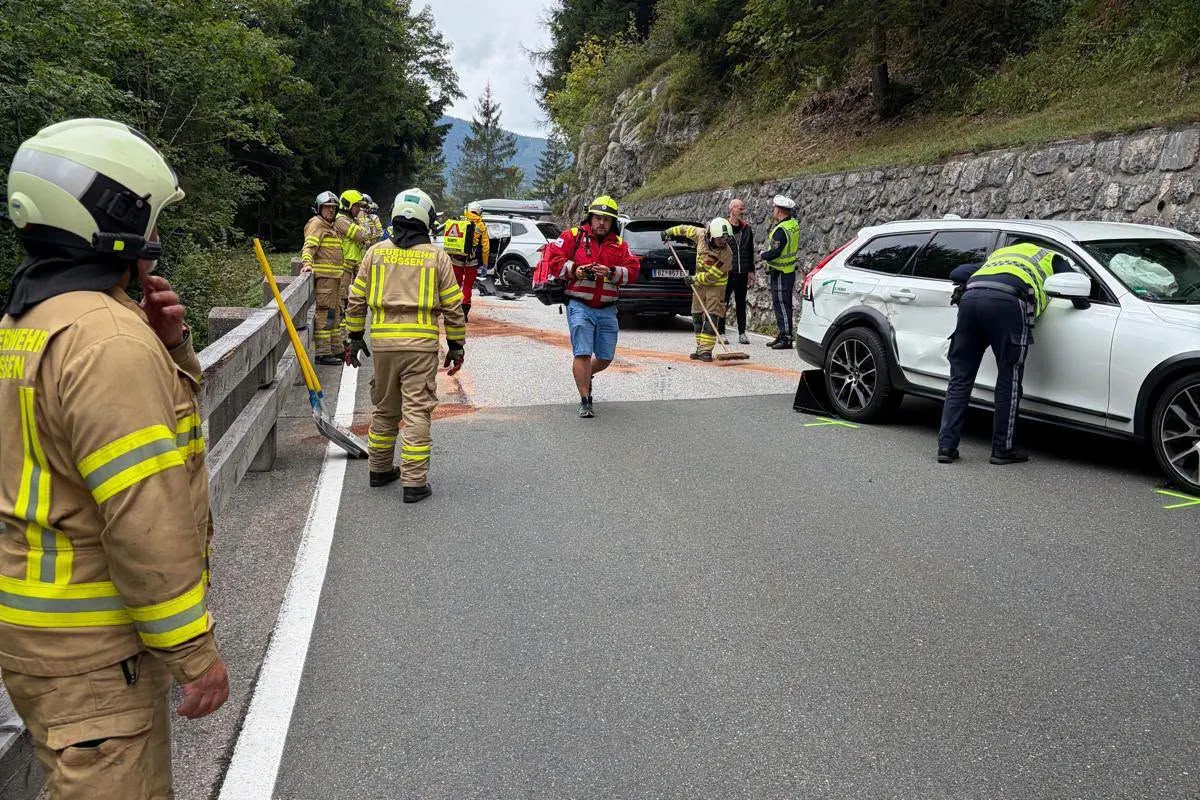 Feuerwehrleute in gelber Schutzkleidung reinigen die Straße, während Polizisten Unfallfahrzeuge inspizieren. Ein weißes Auto zeigt Schäden.